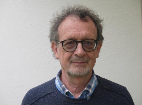 Werner Wintersteiner. &Ouml;sterreichischer Germanist, Friedensp&auml;dagoge, Herausgeber und Hochschullehrer - Mann mit lockigem Haar und Brille, tr&auml;gt einen blauen Pullover und ein kariertes Hemd, steht vor einer hellen Wand.