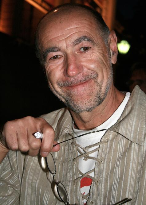 Der &ouml;sterreichischen Musiker Wilhelm 'Willi' Resetarits (Wien 2007) - Ein l&auml;chelnder Mann h&auml;lt eine Brille in der Hand. Er tr&auml;gt ein lockeres, gestreiftes Hemd und hat einen grauen Bart. Hintergrund ist unscharf.